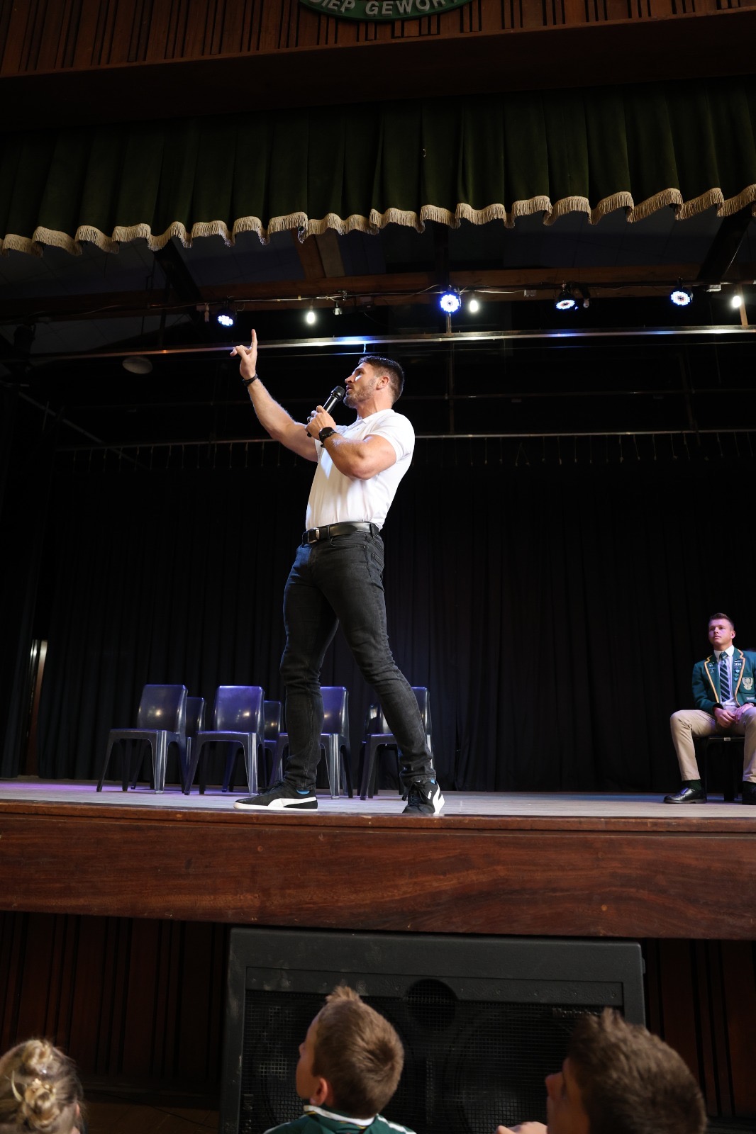 Jaco Kriel on stage pointing upward while speaking into a microphone to a school audience