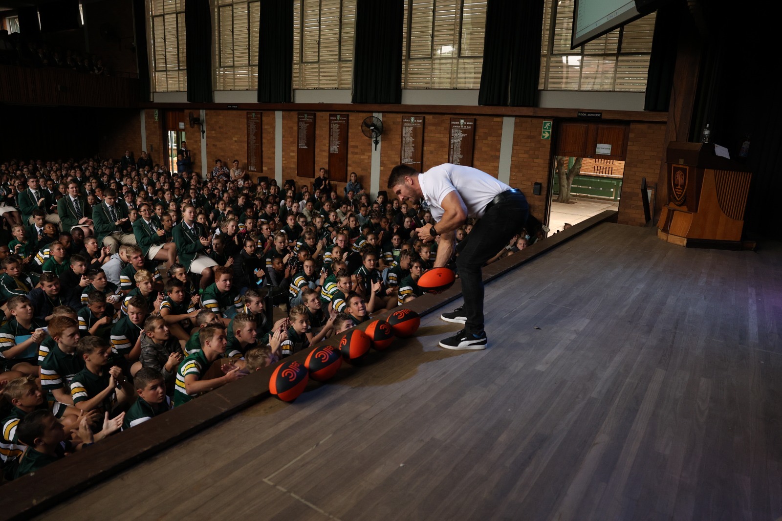 Jaco Kriel rolling a row of signed Cell C rugby balls along the stage edge towards a packed school hall of learners in green blazers
