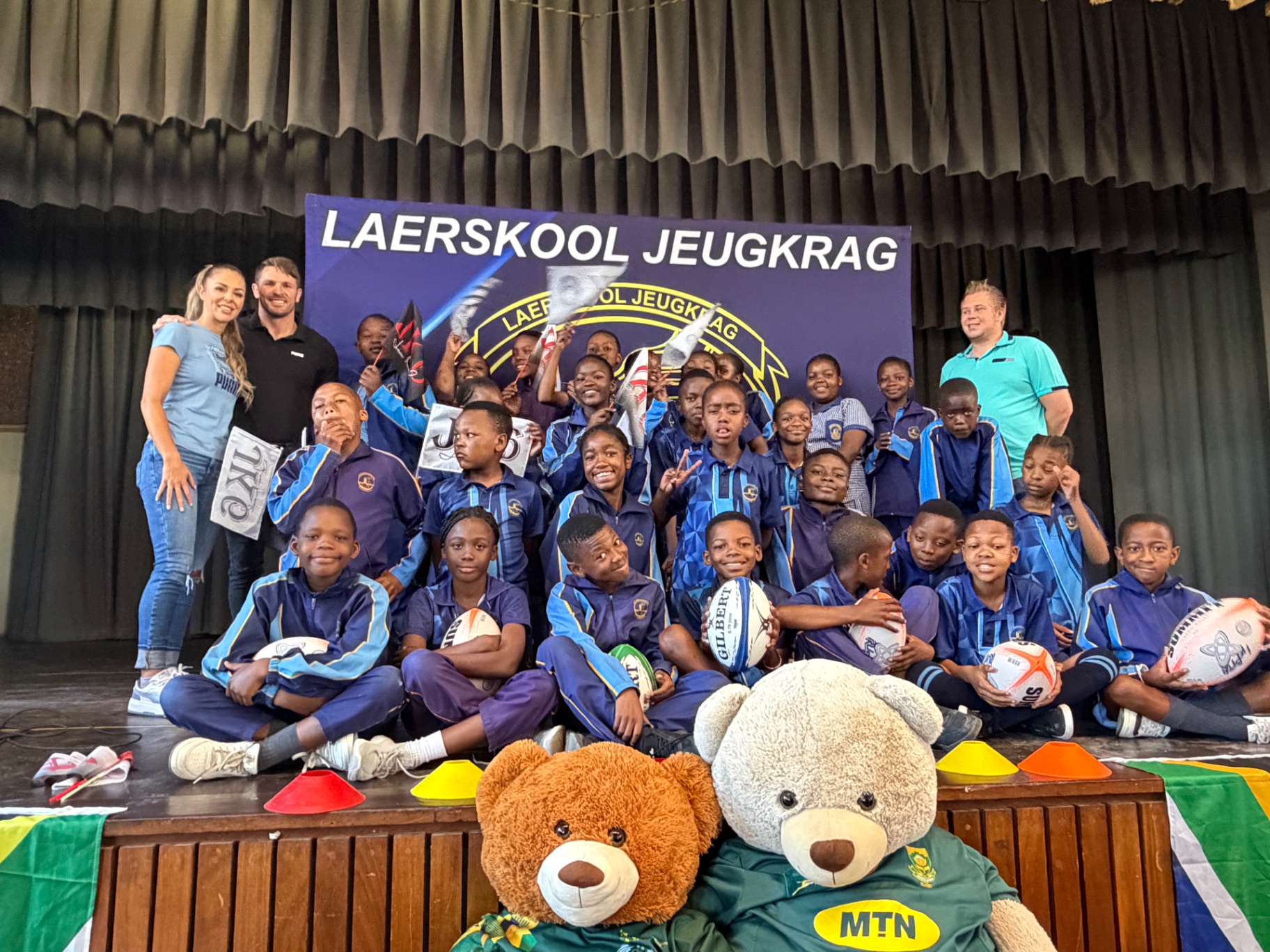 Jaco Kriel and Nastassja Ras with Laerskool Jeugkrag learners holding rugby balls and JK6 flags on stage